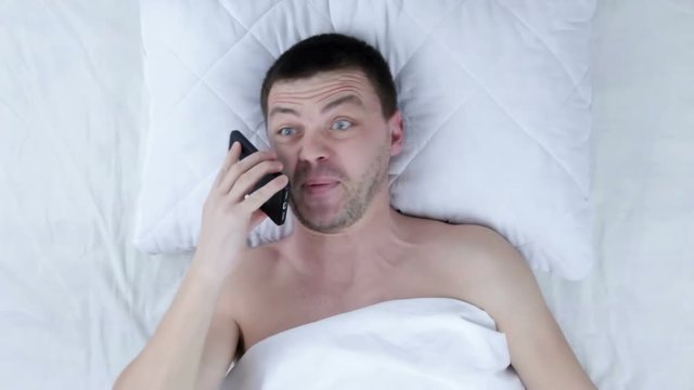 Angry man screaming at phone while lying on bed at midnight
