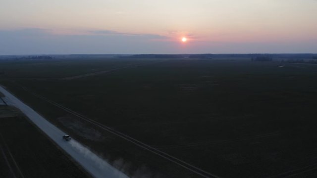 Aerial shot of a car driving on a dirt road, leaving dust behind, during a beautiful sunset
