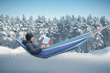 A man reads a red book in a hammock in the winter forest. Mug with a hot drink in his hand