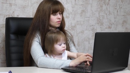 Working mother with her little daughter at table. Busy woman working on laptop with baby on hands. Working mom with beautiful infant on hands in cozy home. Female freelance work. Modern motherhood