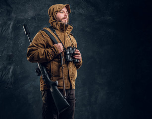 Fototapeta na wymiar Hunter with shotgun holding binoculars and looking sideways. Studio photo against dark wall background