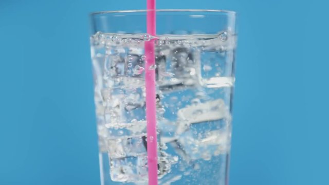 Somebody mix cold water drink with pink drinking straw. Glass full of fizzy with ice cubes. Sparkling soda on blue background, moving bubbles slow motion. Summer party
