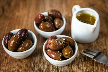Greek Kalamata black red olives in a small white bowls with spices and small bowl with olive oil on wooden table