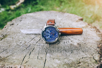 Bid elegant men's watch with old rusty knife is lying on the tree truck in the forest. Accessories for stylish fashion background.