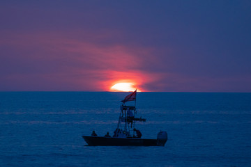 Closeup of a boat on the ocean in front of a glowing setting sun amid the blue and purple clouds of evening, as seen from a beach on the Gulf of Mexico near Englewood, Florida, USA, in early spring