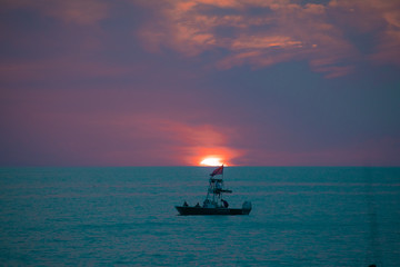 A boat on the teal green ocean water in front of the setting sun amid the blue and purple clouds of evening, as seen from a beach on the Gulf of Mexico near Englewood, Florida, USA, in early spring