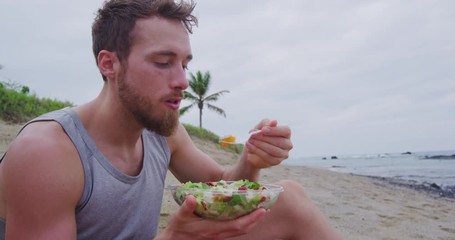 Fitness man eating healthy salad meal at workout. Handsome young muscular male adult sitting on the beach after running workout for lunch break with fresh prepared to go vegan raw vegetables food.