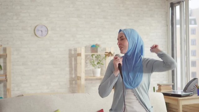 cheerful expressive Muslim girl in hijab in karaoke microphone at home