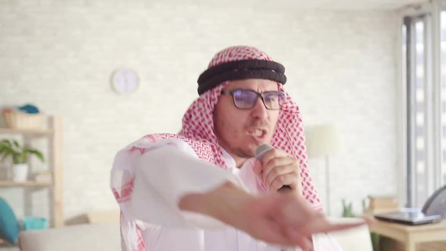 portrait of cheerful Arab man in traditional clothes singing in karaoke microphone at home
