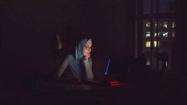 Muslim girl with glasses and a hijab with notebook in the dark search on the Internet at home