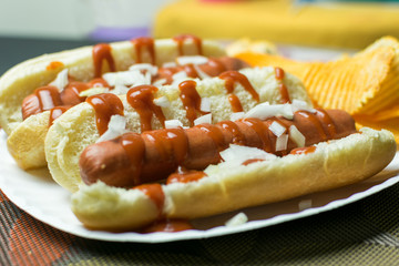Two New York style hot dogs with ketchup, mustard, onions, and a side of potato chips, served on a paper plate