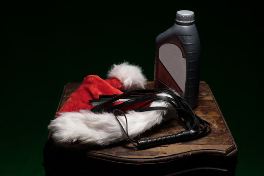 Fuzzy red and white Santa hat, a black whip, and a bottle of motor oil, on an old wooden table, spreading a special kind of holiday cheer