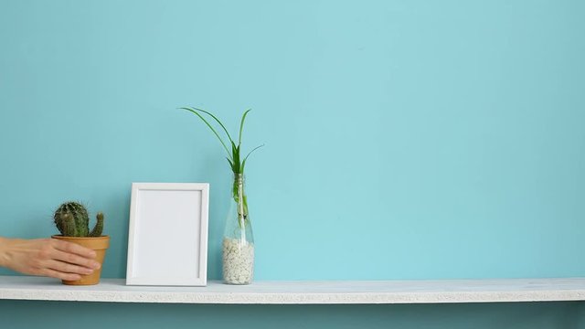 Modern room decoration with Picture frame mockup. White shelf against pastel turquoise wall with spider plant cuttings in water and hand putting down cactus.