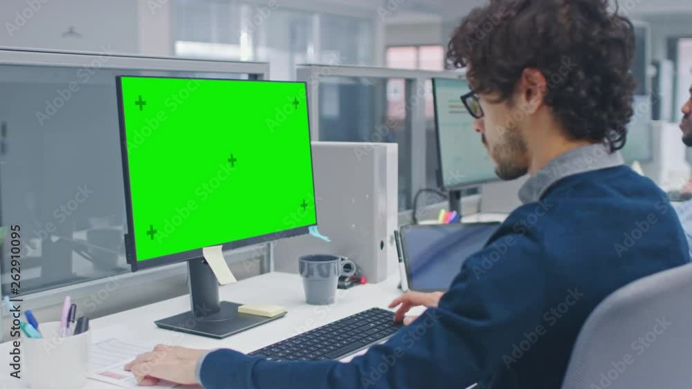 Poster Young Handsome Manager with Curly Hair Works on a Desktop Computer with Green Screen Mock Up. Diverse and Motivated Business People Work on Computers in Modern Open Office.