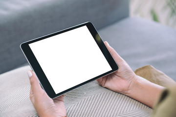 Mockup image of a woman holding black tablet pc with blank white desktop screen  while sitting in living room with feeling relaxed