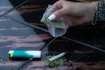 a female hand holds a sachet with a weed, a lighter and a narcotic cigarette in the background