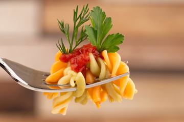 Colored pasta on a fork with tomato sauce and parsley leaf close-up on a blurred wooden background