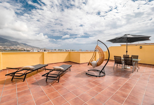 Huge  sunny terrace of a summer holiday property with hanging chair, table, umbrella, sun beds, bamboo sofas and sea view.
