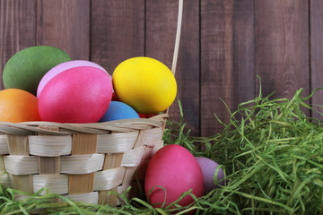 Painted eggs in basket with dry green grass against brown wooden wall. Easter background or egg hunt concept. Pink, yellow, green, orange eggs, hay, side view, close-up