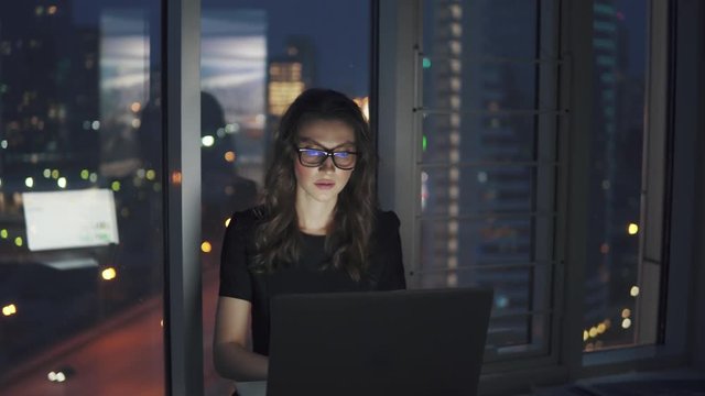 young business woman working late in the office. portrait of a girl with glasses on the background of the night city