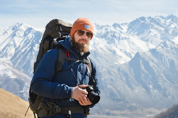 Portrait of a bearded male photographer in sunglasses and a warm jacket with a backpack on his back and a reflex camera in his hands against the background of snow-capped mountains on a sunny day