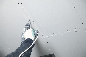 Worker with a drill screwdriver twists the screw into the drywall.