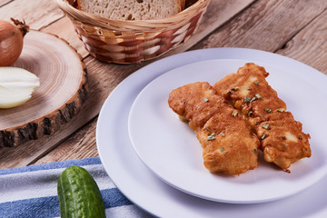 Deep-fried battered cod fillets, in a white plate next to onions, cucumber and bread on a wooden background