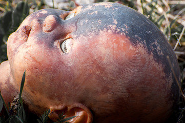 Creepy old doll head. Broken abandoned damaged scary toy in a garbage dump close-up. In the toy, the eye and mouth are damaged. The concept of fear, horror, crime. Very scary.
