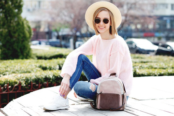 Image of traveling smiling woman walking in spring city. Hipster outfit. Oversize pink sweater, straw hat, round sunglasses and stylish accessories.