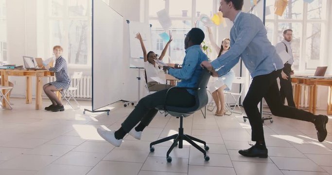 Slow motion crazy fun young black businessman riding office chair, celebrating success with team throwing paper in air.