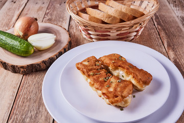 Deep-fried battered cod fillets, in a white plate next to onions, cucumber and bread on a wooden background