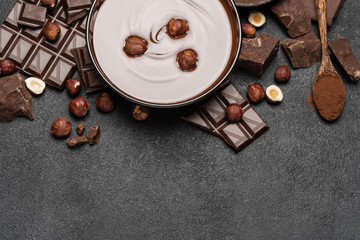 glass bowl of chocolate cream or melted chocolate, pieces of chocolate and hazelnuts on dark concrete background
