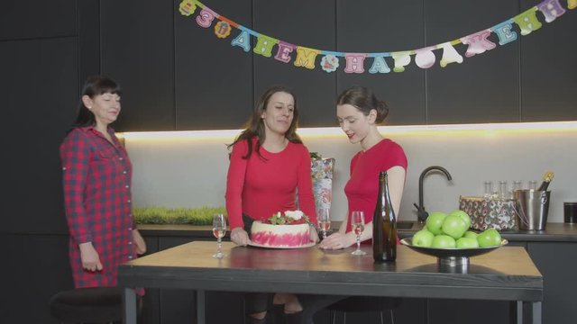 Lovely female receiving congratulations with thirtieth anniversary by cellphone while mother and sister bringing birthday cake to kitchen. Joyful family members kissing, congratulating touched woman.