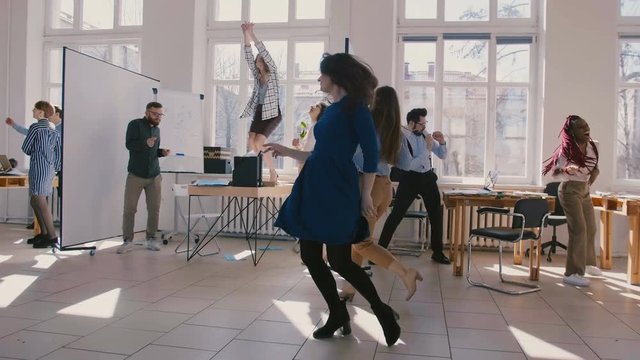 Two young fun female office colleagues dancing together with multiethnic company staff celebrating success slow motion.