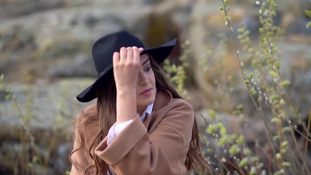 close up of a Beautiful girl in a black hat sits on a large stone. Elegant girl thoughtfully look at distance and straightens hat. Slow motion.