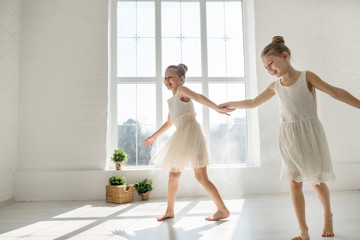 Two little girls in lace dresses like a ballet dancers play and dance and jump near large panoramic window
