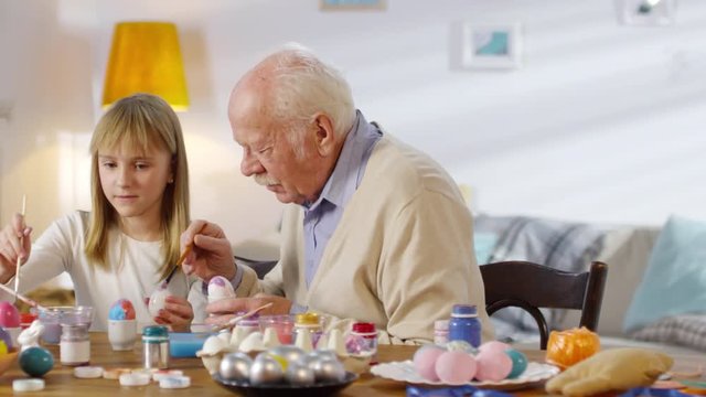 Portrait of cute girl of primary school age and her grandfather painting Easter eggs at home, looking at camera and smiling, tracking shot