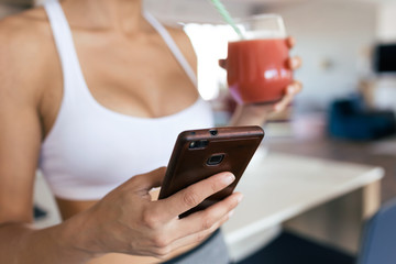 Sporty young woman using her mobile phone while drinking strawberry smoothie in the kitchen at home.
