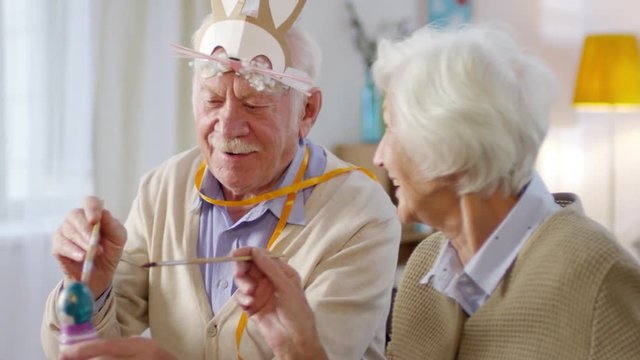 Handheld medium shot of cheerful retired couple painting Easter eggs together, man wearing bunny headband