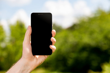 Male hand holding mobile phone with blank black screen outdoors at blurred green park background.