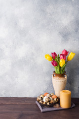 Concept Easter, tulips in a vase and a basket of eggs, a candle on a gray background. Copy space for product or text.
