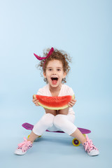 Portrait of funny little curly girl with wide open mouth, holding in hands a slice a watermelon, seated on a skateboards, isolated on a light blue background, with copy space. Expression concept.
