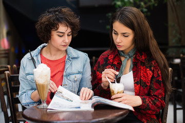 Hipster girls reading magazine at the bar. Girlfriends having a coffee and reading a newspapers at coffee shop. Beautiful girls drinking coffee having a conversation