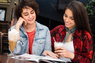 Hipster girls reading magazine at the bar. Girlfriends having a coffee and reading a newspapers at coffee shop. Beautiful girls drinking coffee having a conversation