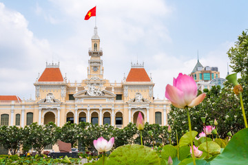 Saigon City Hall with pink lotus flowers (blurred) and blooming plumeria trees in the foreground....