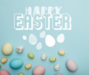top view of painted multicolored eggs scattered and decorative white bunny on blue background with happy Easter lettering