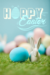 blue painted egg near decorative rabbit on green grass with happy Easter lettering above
