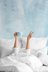 Young woman with cup of hot beverage showing victory gesture in bed - obrazy, fototapety, plakaty