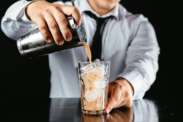Barman makes an orange cocktail. Orange cocktail on a black counter, black background. Cocktail poured from a shaker. 