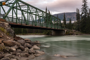 A green iron metal bridge across the Athabasca River in Jasper National Park, waters are smooth due to long exposure